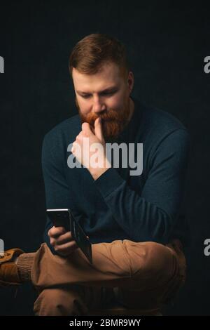 Un giovane ragazzo con una barba rossa di aspetto irlandese si siede e pensa mentre legge un telefono su uno sfondo scuro. Il concetto dell'impatto della rete sociale Foto Stock