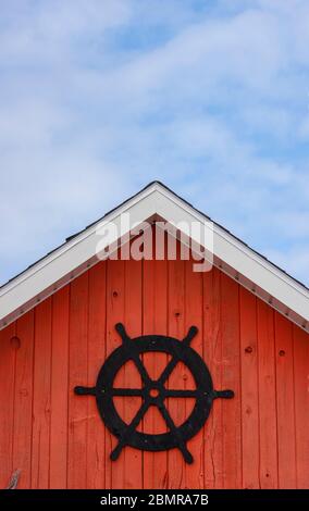 La baracca del pescatore façade. Ruota della nave nera su un bordo dipinto di rosso. Fascia bianca a tegola contro un cielo blu nuvoloso. North rustico Harbour, PEI Canada Foto Stock