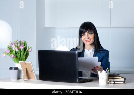 Bella ragazza, brunetta, in un vestito alla sua scrivania tiene carta in mano, sorridendo e guardando la macchina fotografica. Concetto di lavoro per ufficio, donna d'affari, manag Foto Stock