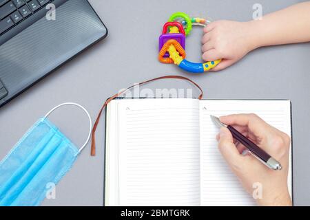 Vista dall'alto della mano dell'uomo che scrive sul notebook mentre la mano del bambino tiene il giocattolo del sonaglino con la maschera medica protettiva e il laptop su sfondo grigio. Lavoro da casa. Foto Stock