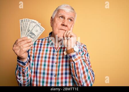 Senior bell'uomo di hoary che tiene mazzo di banconote dollari su sfondo giallo serio faccia pensare alla domanda, molto confuso idea Foto Stock