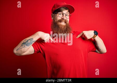 Giovane uomo di consegna bello che indossa gli occhiali e cappuccio rosso su sfondo isolato guardando fiducioso con il sorriso sul viso, puntandosi con le dita p Foto Stock