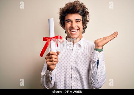 Giovane bell'uomo studente che tiene il diploma in piedi su sfondo rosa isolato molto felice ed eccitato, espressione vincitore che celebra la vittoria urlare Foto Stock