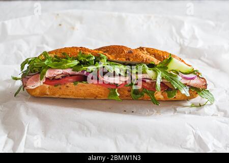 Baguette sandwich con carne, pomodoro, cetrioli e rucola su carta bianca. Cibo da asporto. Foto Stock