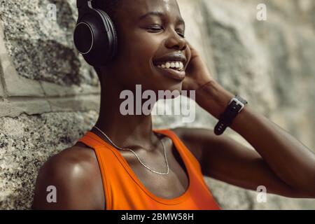 Primo piano di una donna che ascolta musica mentre si fa una pausa all'aperto dopo una sessione di allenamento. Atleta femminile con cuffie rilassanti a parete a poppa Foto Stock