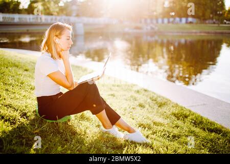 Ragazza che lavora al computer sitingon l'erba in una maschera medica protettiva sul suo viso. Concetto di quarantena indipendente. Foto Stock