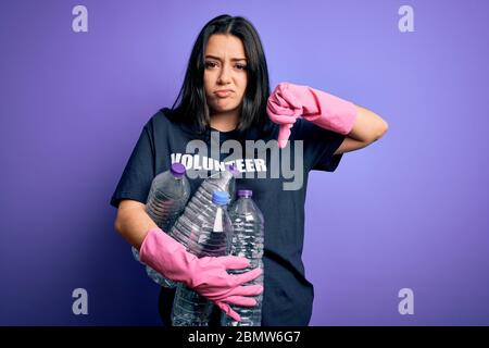 Giovane donna brunetta che indossa volontario t-shirt raccolta bottiglie di plastica su sfondo viola con faccia arrabbiata, segno negativo che mostra dislike con il tum Foto Stock