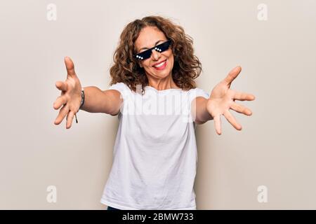 Media età bella donna che indossa divertente vita abbraccio occhiali da sole su sfondo bianco guardando la fotocamera sorridente con le braccia aperte per abbraccio. Allegro Foto Stock