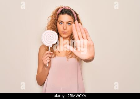 Giovane bella donna bionda con occhi blu mangiare caramella dolce lollipop su sfondo rosa con mano aperta facendo stop segno con serio e fiducioso Foto Stock