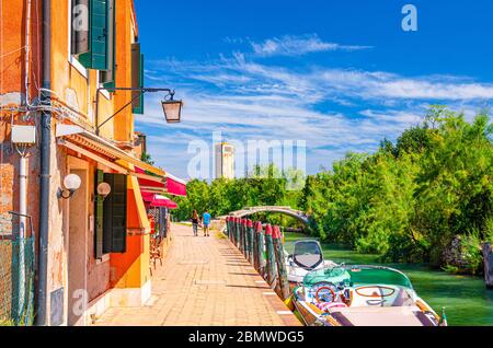 Isola di Torcello con argine lungo il canale d'acqua con barche a motore ed edifici lungo il lungomare. Ponte del Diavolo di pietra, alberi verdi e sfondo torre. Laguna Veneziana, Regione Veneto, Italia. Foto Stock
