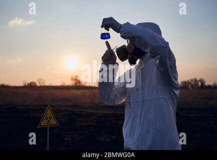 Ambientalista in tuta, maschera a gas, guanti sterili per attività di ricerca sul campo con erba bruciata, tubo di prova con liquido blu, studio di campioni di terreno su territorio bruciato con cartello a rischio biologico. Foto Stock