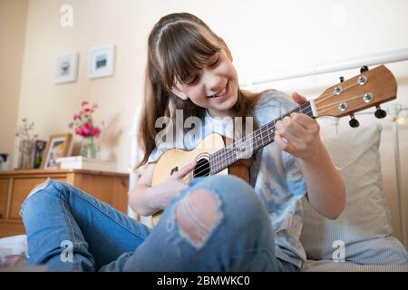 Ragazza che imparano a giocare Ukulele seduto sul letto in camera da letto Foto Stock