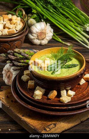 Zuppa di asparagi cremosi purè in ciotola di vetro su piatto nero contro asparagi freschi crudi e verde su sfondo rustico di legno Foto Stock