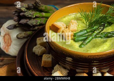 Zuppa di asparagi cremosi purè in ciotola di vetro su piatto nero contro asparagi freschi crudi e verde su sfondo rustico di legno Foto Stock