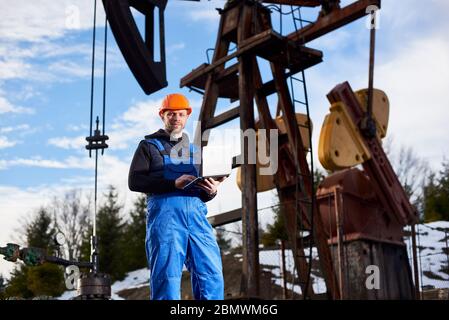 Ritratto di ingegnere petrolo in tute blu e casco arancione, con laptop, in piedi con la schiena a un carro di perforazione olio, controllo unità di pompaggio olio, prendere appunti nel suo computer, sorridendo alla macchina fotografica. Foto Stock