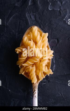 Cucchiaio di posate con cavatappi crudo di grano dorato riccio su sfondo nero scuro testurizzato, vista dall'alto Foto Stock