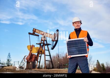 Sorride lavoratore petrolifero in gilet arancione e casco bianco che tiene mini pannello solare in una calda giornata di sole, sullo sfondo il jack pompa e cielo blu, concetto di fonte alternativa di energia Foto Stock