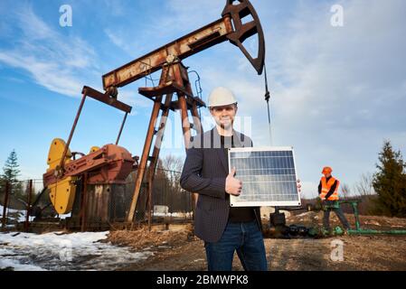 Ritratto di uomo d'affari in giacca, casco, tenendo mini modulo solare, mostrando pollice in su. Operatore di oli che lavora con martinetto pompa sul territorio del campo di olio sullo sfondo. Concetto di energia alternativa Foto Stock