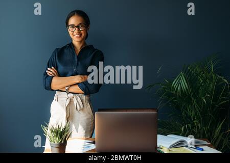 Donna sorridente con le braccia incrociate si trova sul suo posto di lavoro in ufficio Foto Stock