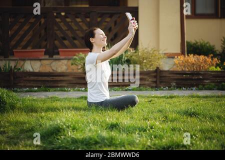 Giovane donna sorridente che fa selfie seduto sull'erba vicino alla casa. Foto Stock