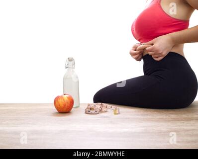 Donna irriconoscibile afferrando le sue maniglie di amore su sfondo bianco con nastro di misurazione, una mela e una bottiglia d'acqua. Concetto di sport a casa. Foto Stock