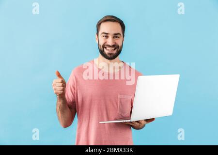 Ritratto di un giovane allegro ed eccitato uomo bearded che indossa la t-shirt in piedi isolato su sfondo blu, tenendo computer portatile, mostrando pollici su ge Foto Stock