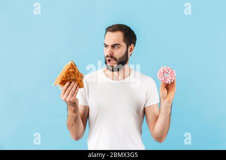 Immagine di giovane dispiaciuto uomo isolato su sfondo blu parete che tiene sandwich e ciambella. Foto Stock