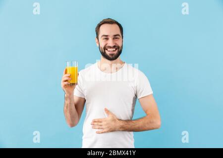 Bell'allegro giovane uomo bearded che indossa la t-shirt in piedi isolato su sfondo blu, mostrando un bicchiere di succo d'arancia fresco Foto Stock