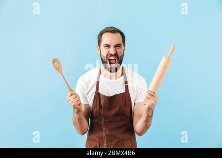 Immagine di giovane uomo chef emotivo urlante isolato su sfondo muro blu con rolling pin e cucchiaio di legno. Foto Stock