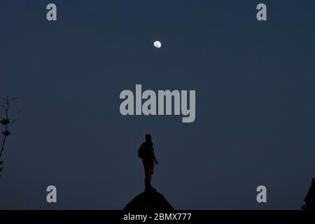 L'uomo si trova retroilluminato su una roccia che guarda fino a una luna quasi piena, con il cielo notturno blu come sfondo Foto Stock