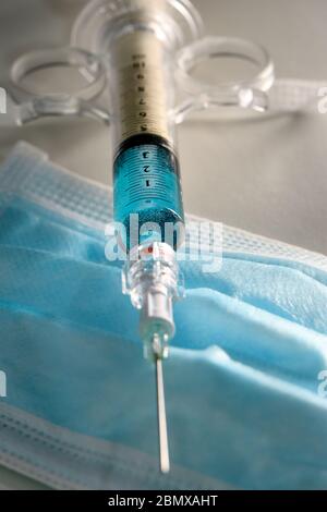 Immagine concettuale di una siringa contenente liquido blu su una maschera facciale blu DPI. Potrebbe essere usato per illustrare la pandemia di covid-19. Foto Stock