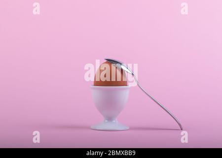 un uovo marrone in una tazza bianca prima di fondo rosa, un cucchiaino da tè sta posando su di esso, primo piano-su Foto Stock