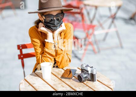 Giovane e triste donna in maschera facciale seduto sulla terrazza del caffè da solo. Concetto di distanza sociale e nuove regole sociali dopo la pandemia di coronavirus. Foto Stock