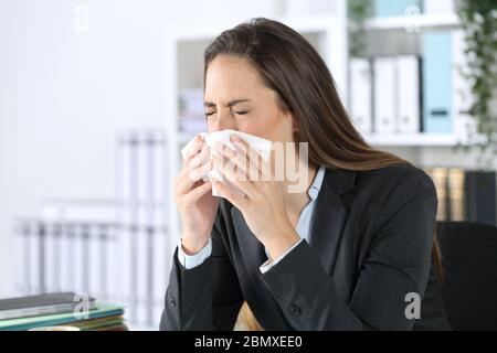 Donna executive starnutita con carta di tessuto che copre la bocca seduto su una scrivania in ufficio Foto Stock