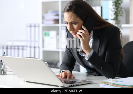 Donna dirigente preoccupata che chiama con uno smartphone per controllare il computer portatile seduto sulla scrivania in ufficio Foto Stock
