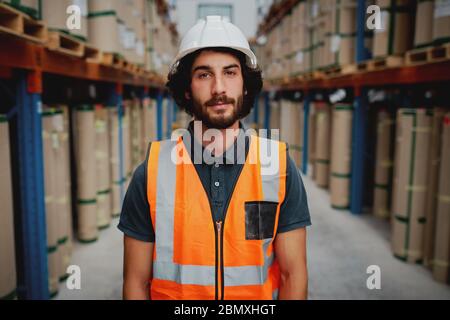 Il responsabile del magazzino indossa una divisa arancione con un casco bianco in piedi nella corsia guardando la telecamera Foto Stock