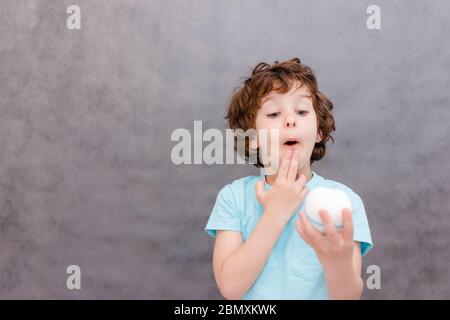 cute ricciolo ragazzo tiene grande dente e sorpreso su sfondo grigio. Il ragazzo perse il primo dente di latte. Concetto di denti sani, clinica odontoiatrica. Foto Stock