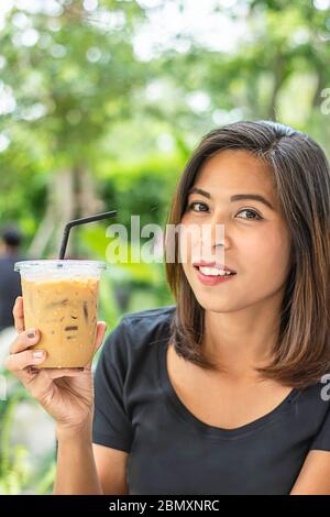 Donna asiatica che tiene un bicchiere di caffè espresso freddo sfondo albero viste sfocate. Foto Stock