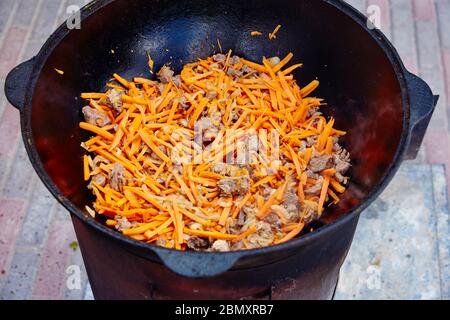 Preparazione di piatto nazionale uzbeko pilaf, pilaw, plov, carota con carne in padella grande. Cottura processo, fuoco aperto. Cottura in un cavolone sul fuoco. Mescolare Foto Stock