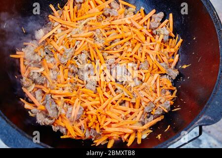 Preparazione di piatto nazionale uzbeko pilaf, pilaw, plov, carota con carne in padella grande. Cottura processo, fuoco aperto. Cottura in un cavolone sul fuoco. Mescolare Foto Stock