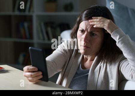 Donna adulta preoccupata che legge le cattive notizie sullo smartphone seduto sul pavimento di notte a casa Foto Stock