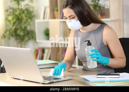 Donna imprenditore con maschera disinfettante tastiera computer con igienizzatore e salvietta seduta su una scrivania a casa Foto Stock