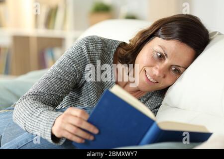 Felice donna di mezza età che legge libro di copertura duro seduta su un divano a casa Foto Stock