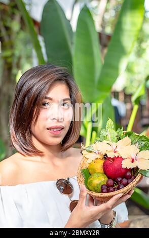 Donna a mano tenendo l'intreccio cesti di bambù con Apple, arancio e uva sfondo sfocato alberi. Foto Stock