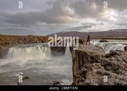 Una donna che si erge su una roccia che si affaccia sulla cascata Godafoss durante il giorno nel nord-est dell'Islanda Foto Stock