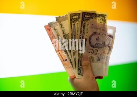 Mano tenendo mix di banconote dollaro americano, rupia indiana, ringgit malese, dollaro di singapore e più di fronte a una bandiera indiana che mostra Foto Stock