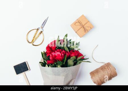 Vista dall'alto del bouquet di peonie avvolto in carta ecologica accanto alla corda di iuta, scatola regalo, forbici e piatto di gesso in legno su sfondo bianco. Foto Stock