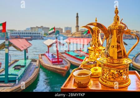 Gustatevi un caffe' caldo dalla tradizionale caffettiera di ottone arabo con dallah sulla riva del Dubai Creek con una vista sulle barche di abra, Dubai, UAE Foto Stock