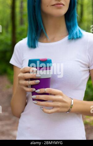 Giovane adulto bella donna con capelli blu in piedi nel giardino esterno tenendo una tazza riutilizzabile con bevanda calda Foto Stock