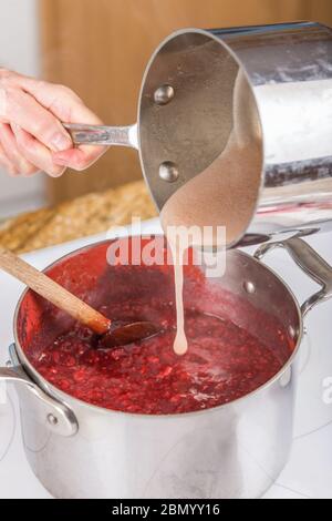 Donna versando di pectina di frutta e di acqua nella miscela di lamponi come una parte di confettura di lamponi. (MR) (PR) Foto Stock